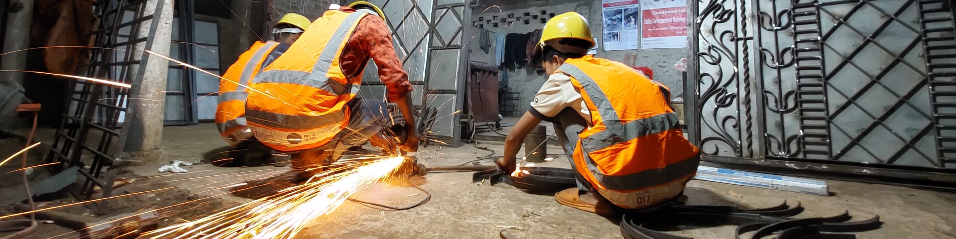 Workers in safety vests and helmets use power tools to cut metal in a workshop, with sparks flying from the cutting process.