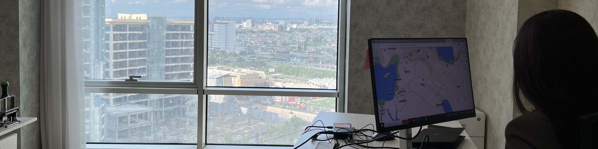 A person working at a desk with a computer displaying a map, overlooking a cityscape through a large window.