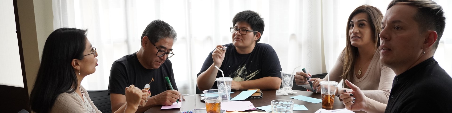 Five people sitting at a round table during an LGBTIQA+ workshop, discussing and making notes.