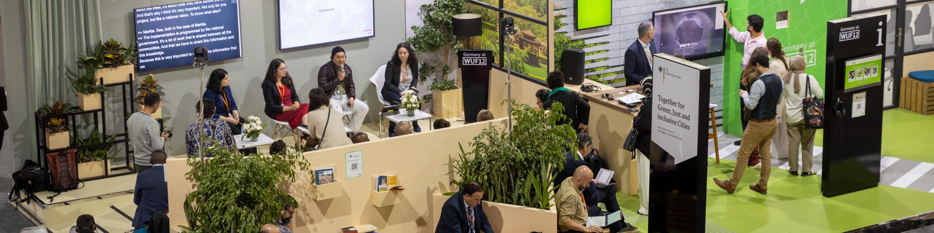 An exhibition booth with many visitors at the World Urban Forum 12. A panel discussion is being held.