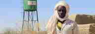 A man – Ibrahim Yatara – standing in front of a green water tank in an arid landscape.