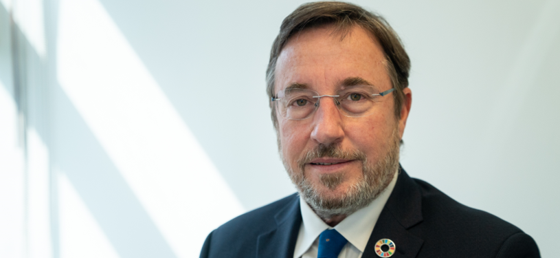 UNDP Director Achim Steiner wearing a suit and tie with a UN goal emblem, smiling at the camera.