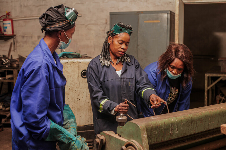 A woman instructs two other women at a metal hand-washing unit.