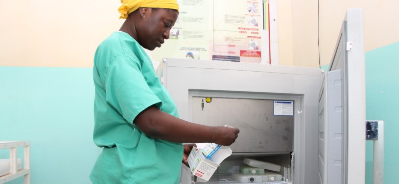 A woman stands in front of an open refrigerator and fills it with medications. She is wearing medical workwear.