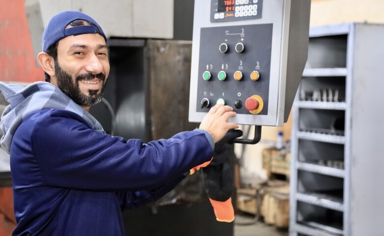 A man wearing a cap and a work jacket smiles as he operates a control panel in an industrial setting.