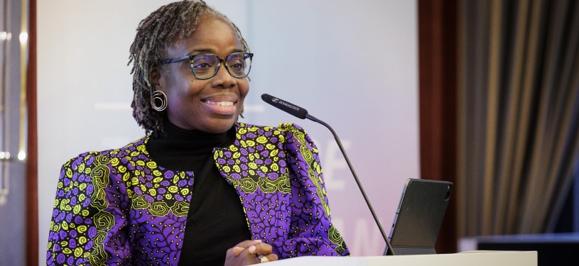 A woman (Mavis Owusu-Gyamfi) in a purple blazer stands at a podium with a microphone and smiles.