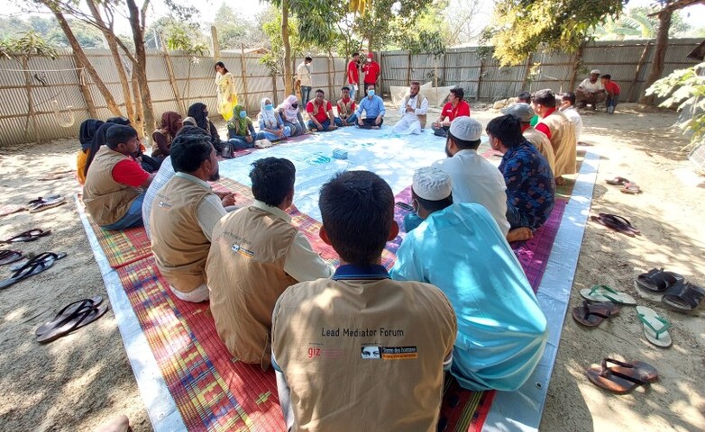 Participants sit in a circle on colorful mats during a meeting of the mediators' forum in a Rohingya camp, held outdoors and surrounded by trees and a fence.