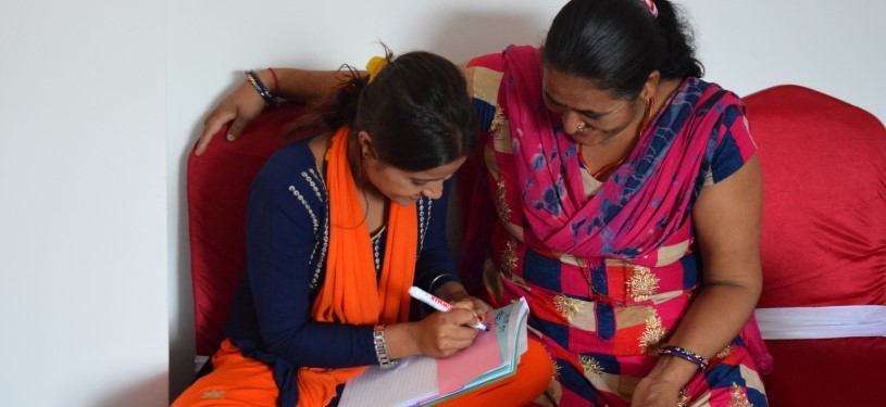 Two women in colorful clothes bend over a notepad together and write something on it.