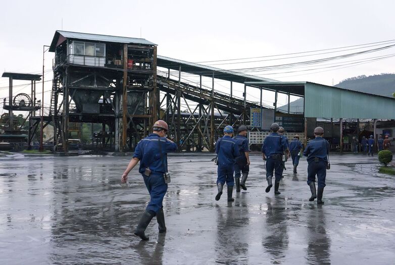 A group of coal workers crosses a rain-soaked farm, with a conveyor belt system in the background.