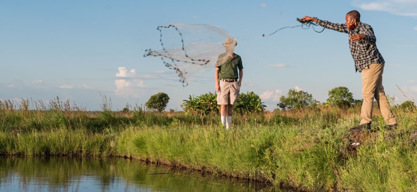 A man at a pond throws out a fishing net; a second man is in the background.