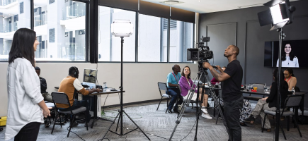 A cameraman takes shots of a woman in a well-lit room while people sit and work nearby.