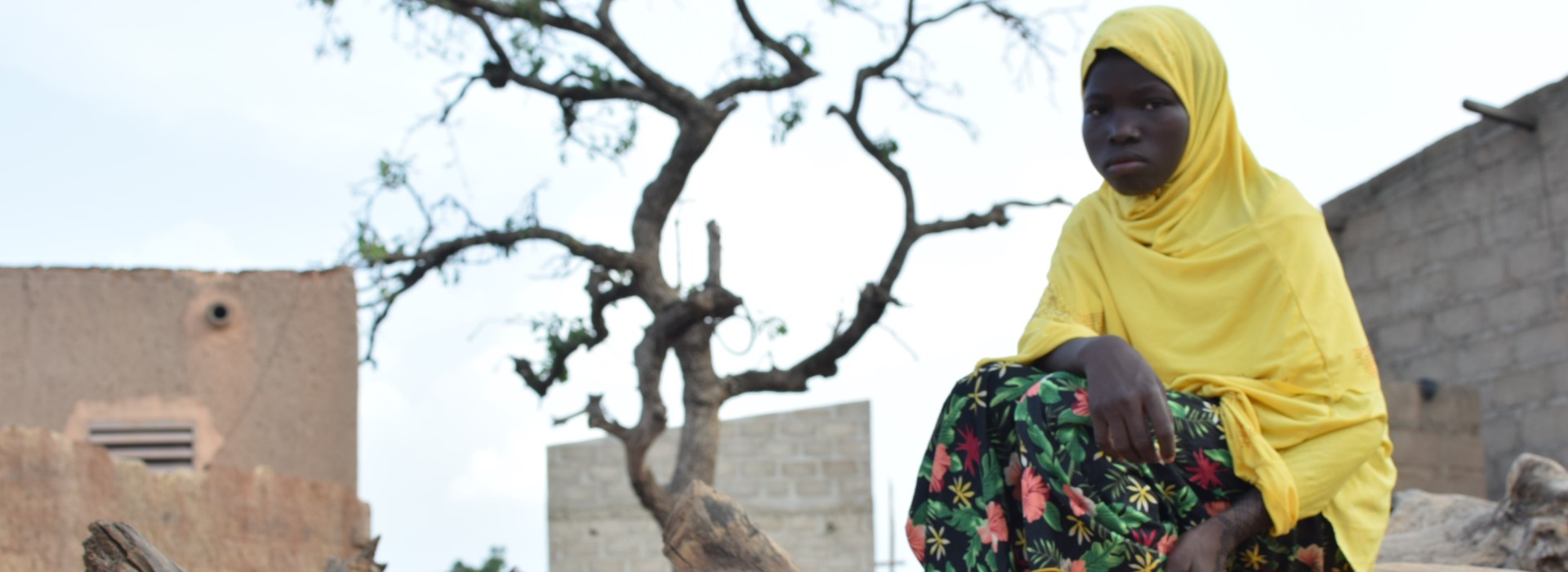 The girls Bouama, wrapped in a yellow scarf, sits on a tree trunk and looks sadly into the camera.