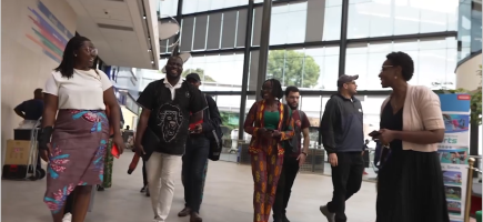  A group of people, including several women and men, walk through a spacious, modern building with large windows, engaged in conversation and smiling.