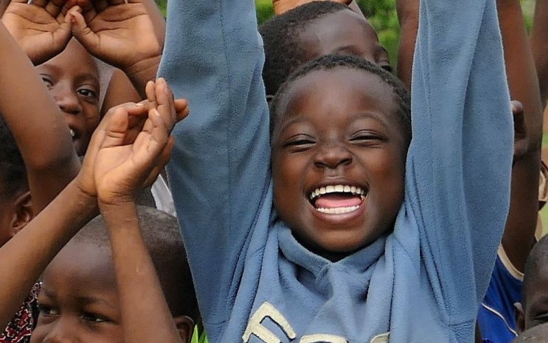 A group of children stretch their hands in the air, all laughing into the camera.