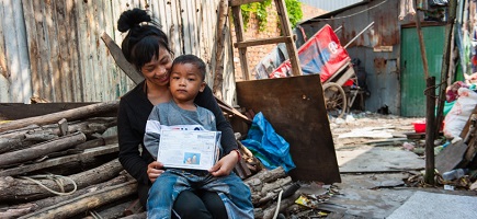 A woman with a small child in her lap holds up to the camera a laminated card featuring her personal details.