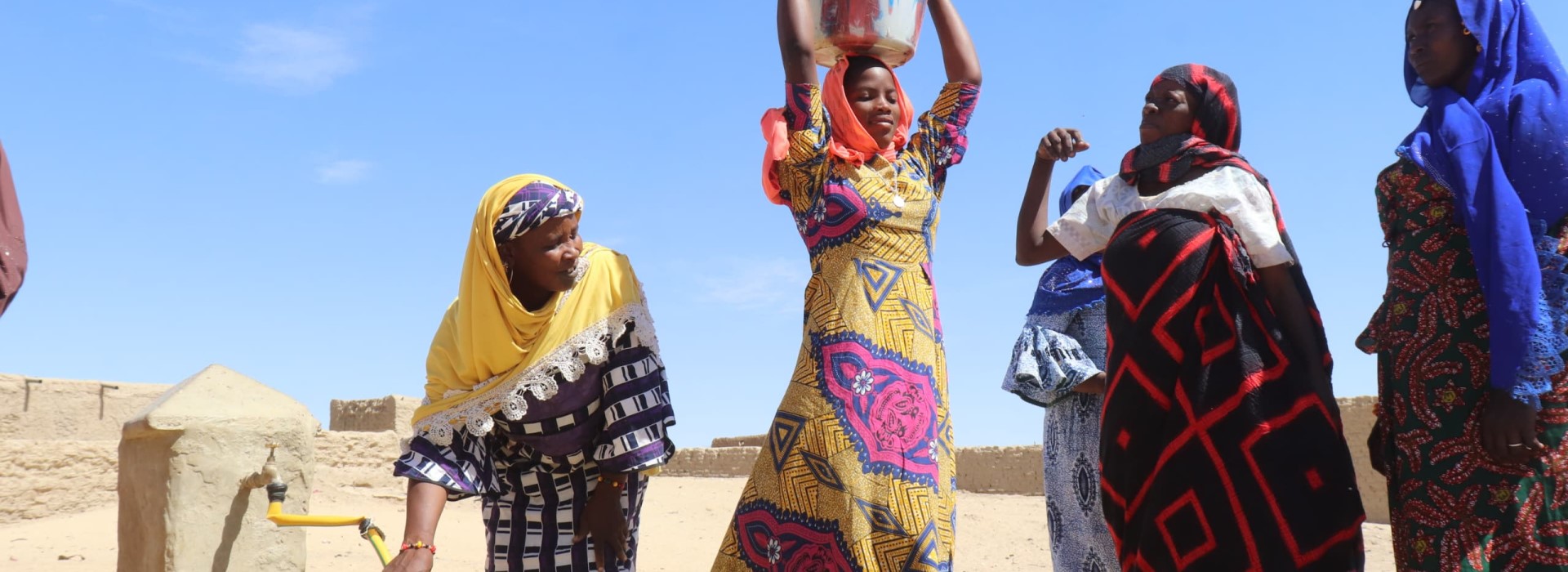 Women in brightly coloured dresses fetching water from a well and carrying it in containers on their heads.