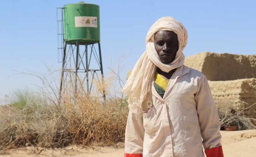 A man – Ibrahim Yatara – standing in front of a green water tank in an arid landscape.