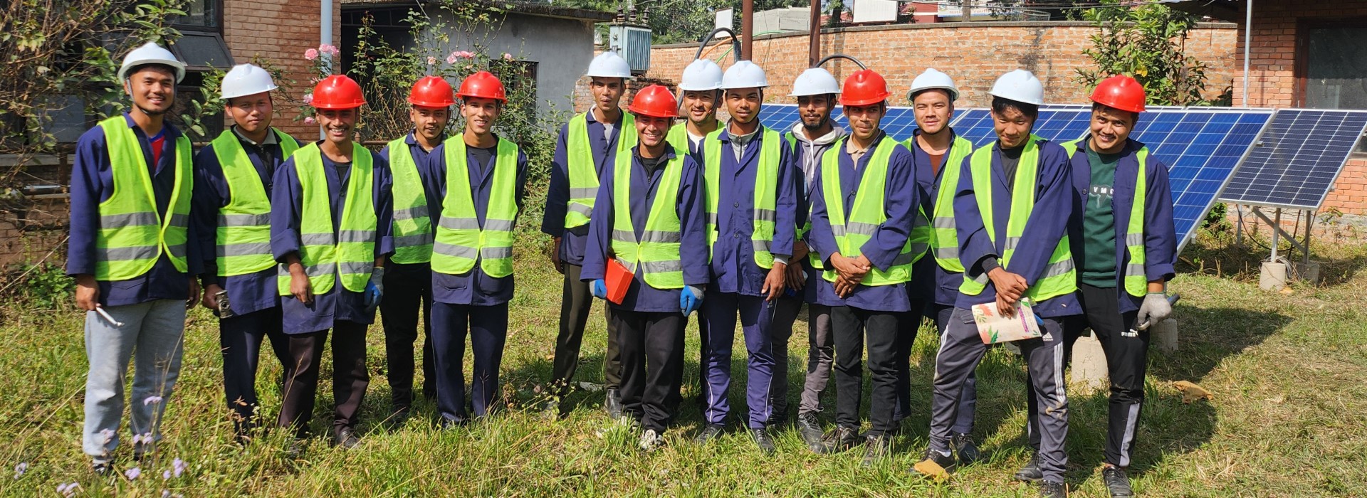 A group photo of 14 men in yellow vests and white and red safety helmets, with two solar panels in the background.