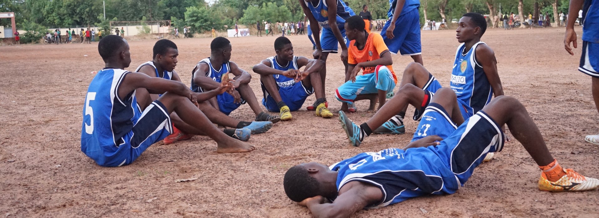 A group of young people dressed in basketball gear sits on the floor as a coach talks to them.