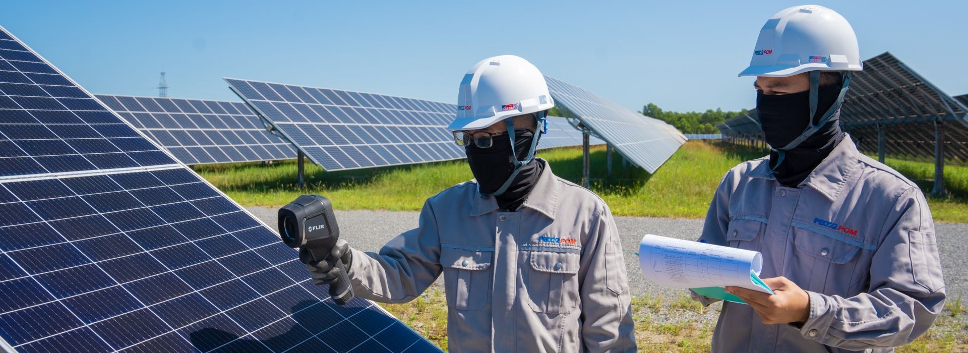 Two men in protective clothing hold a measuring device to a solar panel in a solar park.