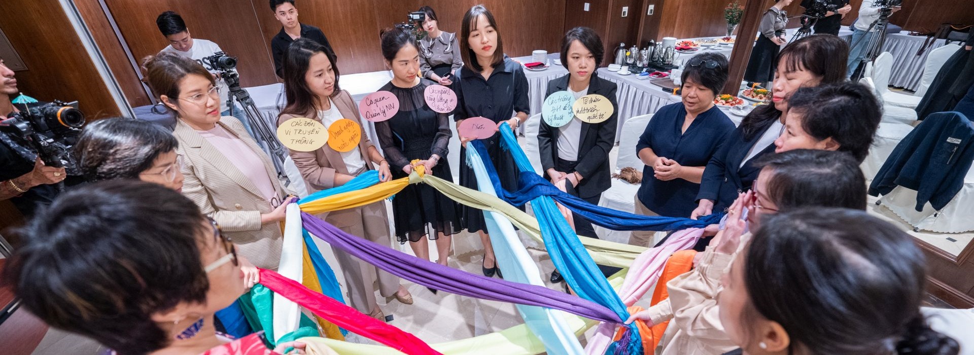 13 businesswomen stand in a circle and hold a net of colorful scarves.