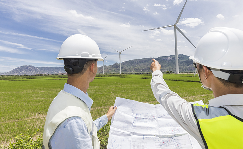 Two men in white construction helmets hold a map in their hands, one points to three wind turbines in the background.